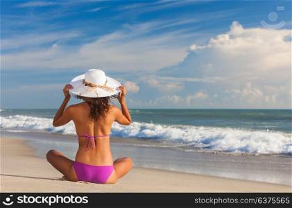 Rear view of beautiful young woman in bikini and a white sun hat sitting on a deserted tropical beach with blue sky
