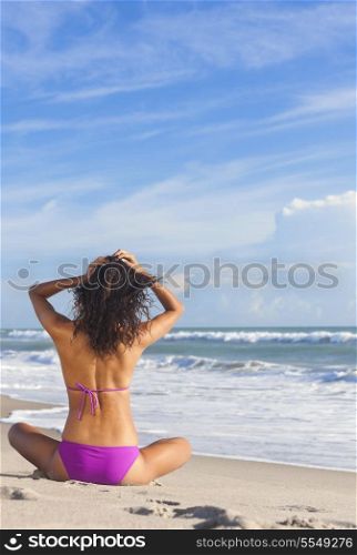 Rear view of beautiful young woman in bikini sitting cross legged on a deserted tropical beach with blue sky