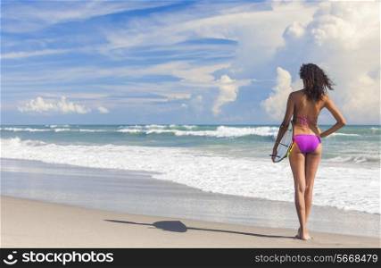 Rear view of beautiful young woman surfer girl in bikini with surfboard standing in the surf on a beach