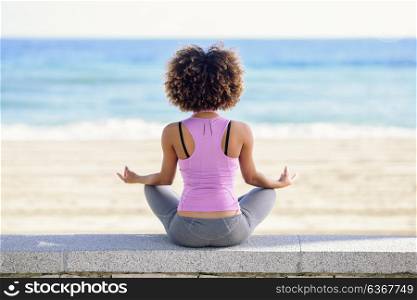 Rear view of black woman, afro hairstyle, doing yoga in the beach. Young Female wearing sport clothes in lotus figure with defocused background.