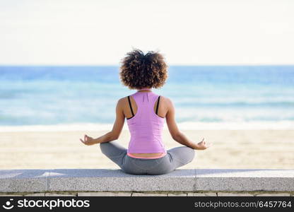 Rear view of black woman, afro hairstyle, doing yoga in the beach. Young Female wearing sport clothes in lotus figure with defocused background.