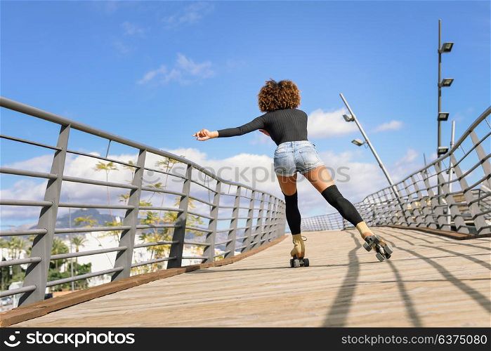 Rear view of black woman, afro hairstyle, on roller skates riding outdoors on urban bridge. Young girl rollerblading on sunny day.