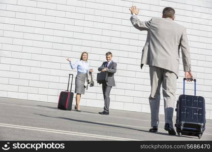 Rear view of businessman carrying luggage waving hand to colleagues