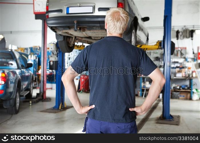Rear view of car repairer looking at car in auto repair shop with hands on waist