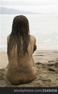 Rear view of Caucasian young adult nude woman sitting on beach.