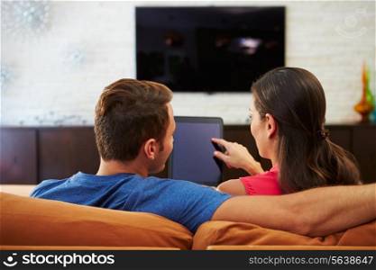 Rear View Of Couple Sitting On Sofa Using Digital Tablet