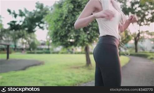 Rear view of fit young slender asian woman stretching body in a green park, with medical mask hanging on arm, getting body ready for work out, cooling down after work out, activity during epidemic