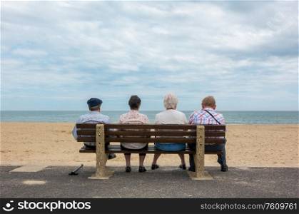 Rear view of four anonymous senior people, old couples, elderly men and women sitting on a bench at a beach