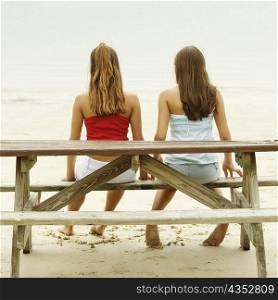 Rear view of girl and a teenage girl sitting on a picnic table on the beach