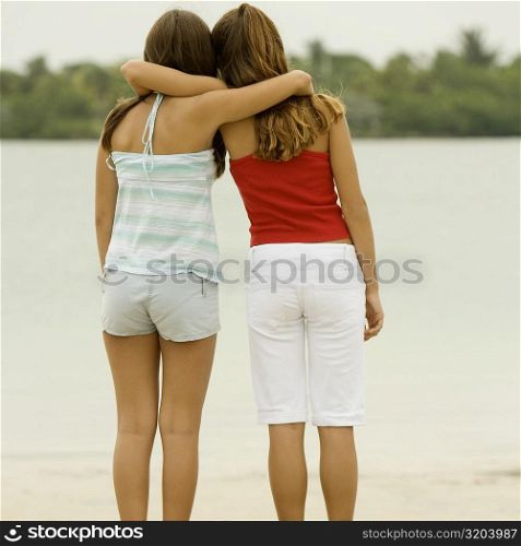 Rear view of girl and a teenage girl standing with their arms around each other on the beach