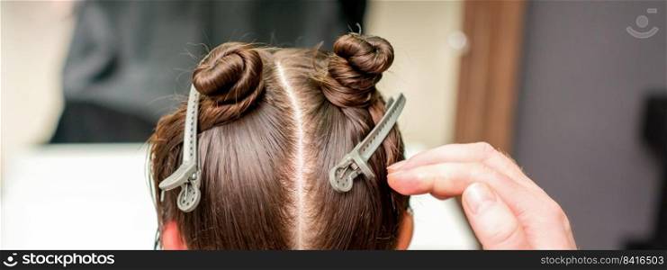 Rear view of hand of hairdresser doing haircut of young woman with hair clips on her hair in hair salon. Hair clips on hair of young woman