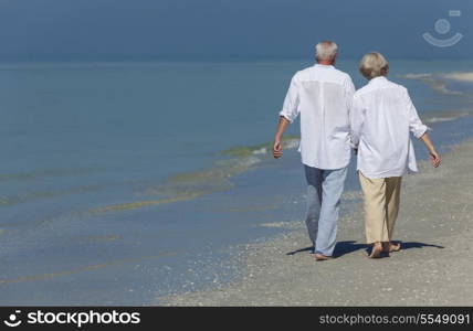 Rear view of happy senior man and woman couple walking and holding hands on a deserted tropical beach with bright clear blue sky