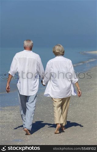 Rear view of happy senior man and woman couple walking and holding hands on a deserted tropical beach with bright clear blue sky