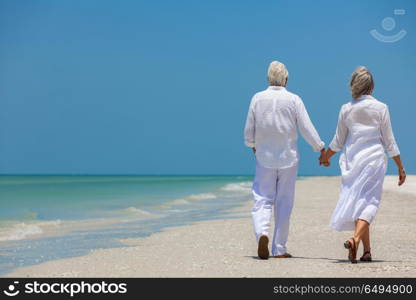 Rear view of happy senior man and woman couple walking and holding hands on a deserted tropical beach with bright clear blue sky. Happy Senior Couple Walking Holding Hands Tropical Beach