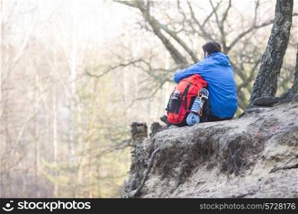 Rear view of hiker with backpack sitting on edge of cliff in forest