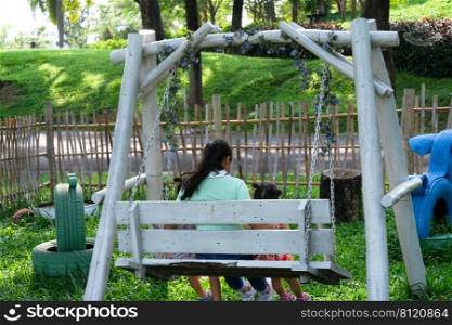 Rear view of little girls sitting on a swing under tree with mother in the summer park. Happy family having fun in the park.