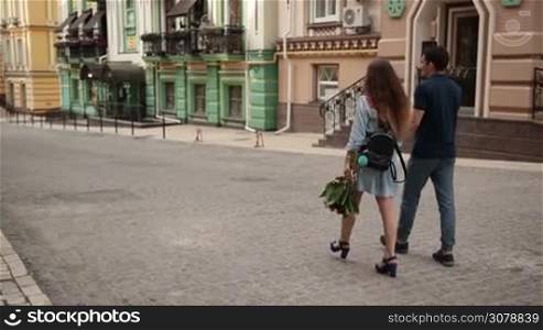 Rear view of loving couple stepping down cobblestone sidewalk during romantic date. Attractive woman carrying bunch of flowers holding hand of boyfriend walking down the street while enjoying time together outdoors. Slow motion. Steadicam stabilized shot.