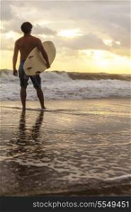 Rear view of male surfer with white surfboard standing on a beach at sunset or sunrise
