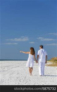 Rear view of man and woman romantic couple in white clothes walking holding hands and pointing on a deserted tropical beach with bright clear blue sky