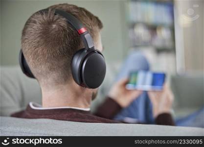 Rear View Of Man Relaxing On Sofa Wearing Wireless Headphones Streaming Film Music Or Podcast From Mobile Phone