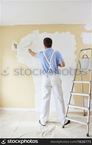 Rear View Of Man Wearing Overalls Painting Wall In Room Of House With Paint Roller