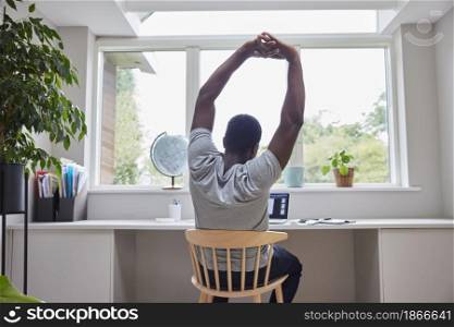 Rear View Of Man Working From Home On Computer In Home Office Stretching At Desk