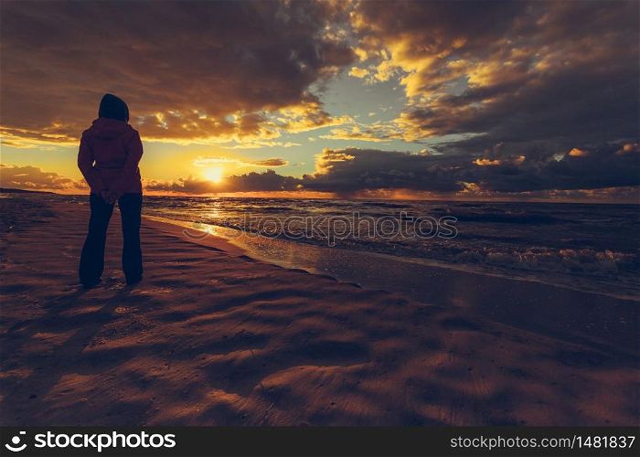 Rear view of mature woman silhouette standing on beach looking at horizon enjoying dramatic sunset in cold weather day. Woman enjoying dramatic sunset on sea