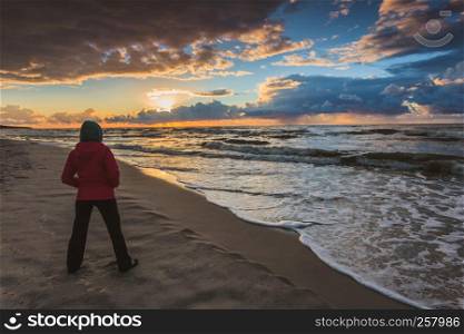 Rear view of mature woman silhouette standing on beach looking at horizon enjoying dramatic sunset in cold weather day. Woman enjoying dramatic sunset on sea