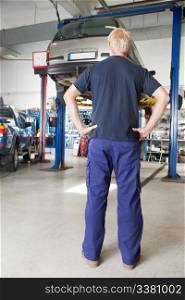 Rear view of mechanic looking at car in auto repair shop with hands on waist