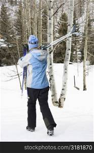 Rear view of mid adult Caucasian female skier wearing blue ski clothing walking and carrying skis on shoulder.