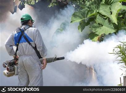 Rear view of outdoor healthcare worker using fogging machine spraying chemical to eliminate mosquitoes and prevent dengue fever on overgrown in slum area