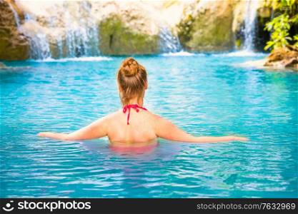 Rear view of red haired young woman in pink bikini swimsuit relaxes in emerald blue water of tropical lake with waterfall. Erawan National park, Kanchanaburi, Thailand