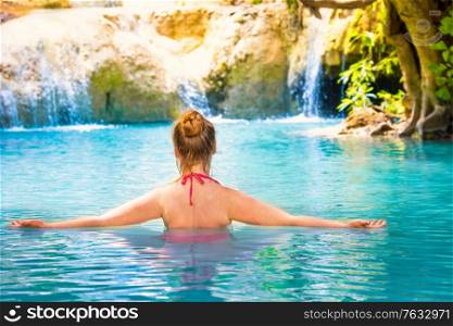 Rear view of red haired young woman in pink bikini swimsuit relaxes in emerald blue water of tropical lake with waterfall. Erawan National park, Kanchanaburi, Thailand