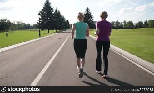 Rear view of senior female joggers running in park road in sportswear over beautiful landscape background. Active fit blonde women jogging during outdoor workout in park. Slow motion. Steadicam stabilized shot.
