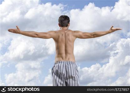 Rear view of shirtless young man standing arms outstretched against cloudy sky