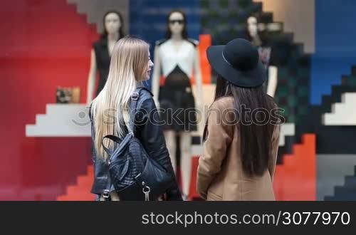 Rear view of shopaholic young women looking at clothing store display outside the shop. Two attractive long hair girlfriends with shopping bags standing in front of clothing window display with with mannequins in fashionable dresses.