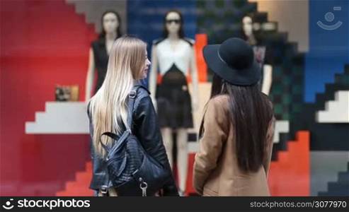 Rear view of shopaholic young women looking at clothing store display outside the shop. Two attractive long hair girlfriends with shopping bags standing in front of clothing window display with with mannequins in fashionable dresses.