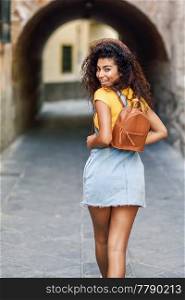 Rear view of smiling black tourist woman with curly hair walking on the street. Arab traveler girl in casual clothes in the street. Happy female wearing yellow t-shirt and denim dress in urban background.
