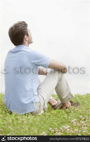 Rear view of thoughtful young man sitting on grass against sky