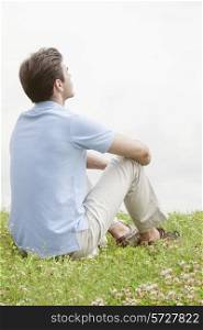 Rear view of thoughtful young man sitting on grass against sky