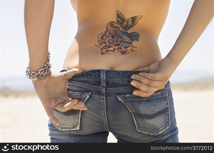 Rear view of torso and derriere of Caucasian mid-adult woman with humminbird tattoo on back and hands tucked in back of jeans.