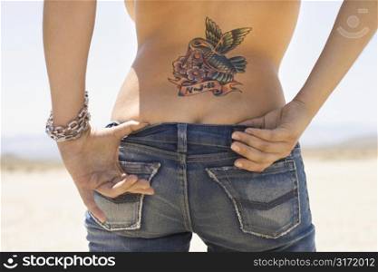Rear view of torso and derriere of Caucasian mid-adult woman with humminbird tattoo on back and hands tucked in back of jeans.
