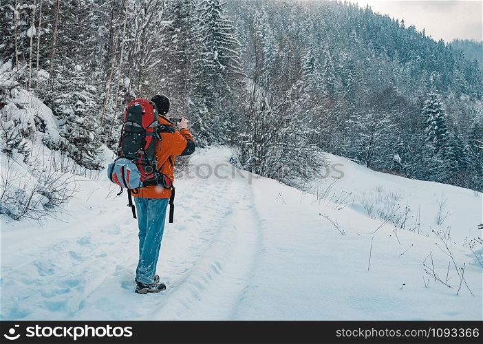 Rear view of tourist taking photo of mountain forest landscape on snowy winter day. Blue jeans, orange garment, red backpack. Hiking travel extreme concept