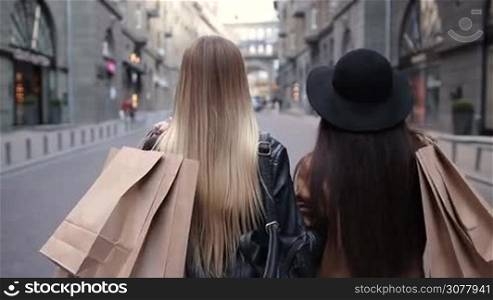 Rear view of two attractive teenage women with shopping bags over theirs shoulders going along city street. Slow motion. Multhiethnic girlfriends carrying shopping bags, walking on sidewalk and looking back with toothy smiles after day shopping.