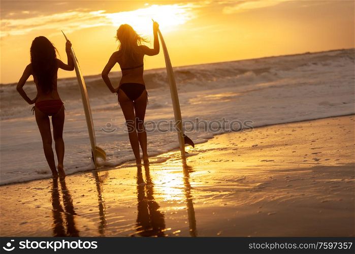 Rear view of two beautiful sexy young women surfer girls in bikinis with white surfboards on a beach at sunset or sunrise