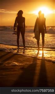Rear view of two beautiful sexy young women surfer girls in bikinis with white surfboards on a beach at sunset or sunrise