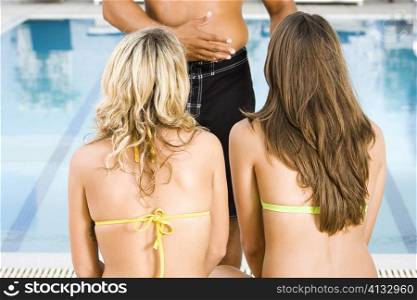 Rear view of two young women listening to a mid adult man at the poolside