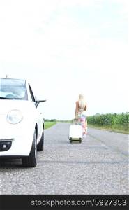 Rear view of woman with luggage leaving broken down car on country road