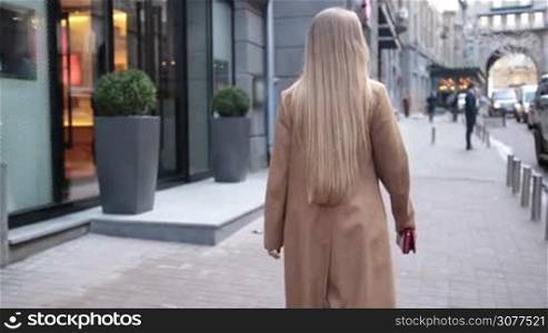 Rear view of young beautiful blonde woman with wonderful long hair wearing elegant camel coat and holding red purse in her hands while walking on city street. Elegant female going for shopping. Slow motion.