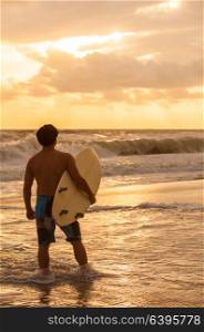 Rear view of young man male surfer with white surfboard looking at surf on a beach at sunset or sunrise